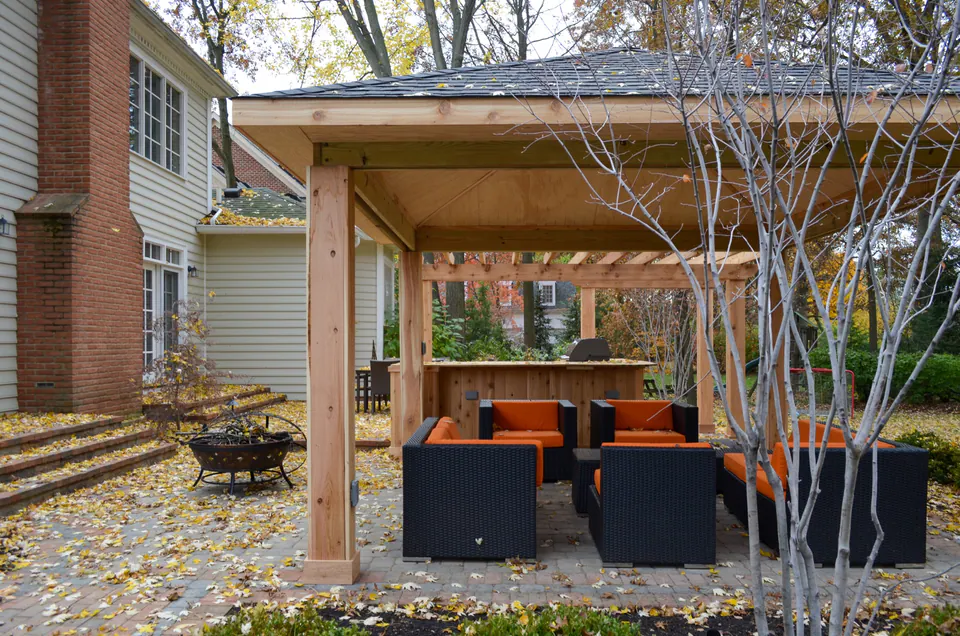 Exterior pavilion view with lounge seating, wood posts, paver patio, leaf-covered beds, and brick house.